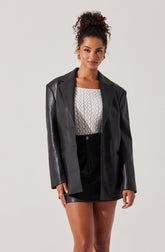 A woman is wearing a black leather blazer with a notched lapel collar, layered over a white textured top with a square neckline. She has paired this with a black high-waisted mini skirt with visible stitching details. She accessorizes with gold hoop earrings and a gold ring on her left hand.